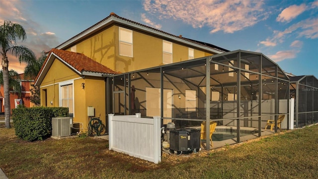 back of property at dusk with stucco siding, central air condition unit, a lawn, a tile roof, and glass enclosure