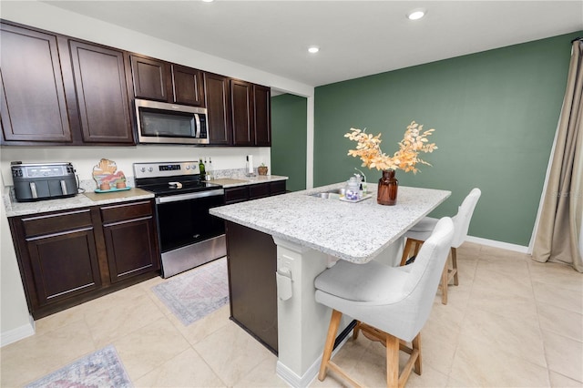 kitchen with a kitchen breakfast bar, an island with sink, dark brown cabinets, light stone counters, and stainless steel appliances