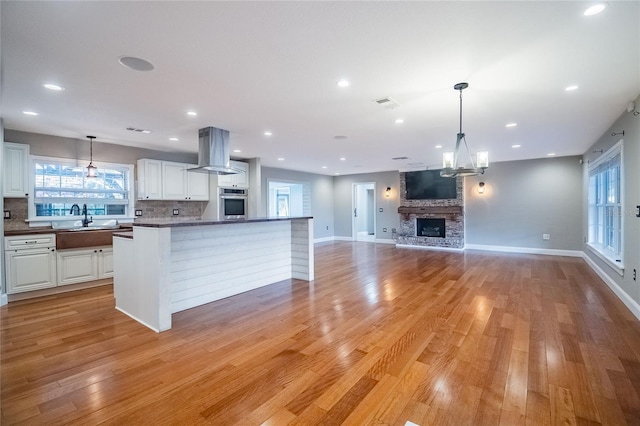 kitchen featuring wall chimney exhaust hood, white cabinetry, light hardwood / wood-style flooring, oven, and a stone fireplace