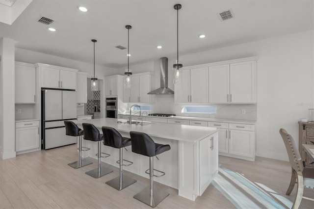 kitchen with wall chimney exhaust hood, a kitchen island with sink, white refrigerator, decorative light fixtures, and white cabinetry