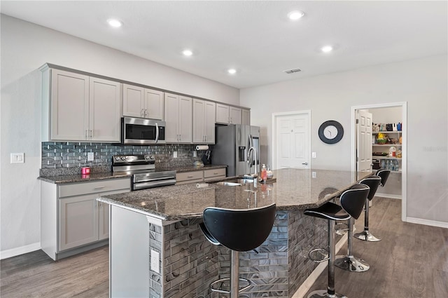 kitchen featuring dark stone counters, a center island with sink, gray cabinets, appliances with stainless steel finishes, and a breakfast bar area