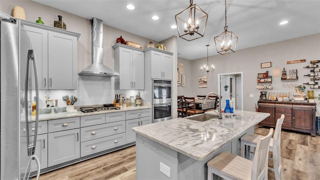 kitchen with appliances with stainless steel finishes, wall chimney exhaust hood, a kitchen island with sink, sink, and hanging light fixtures