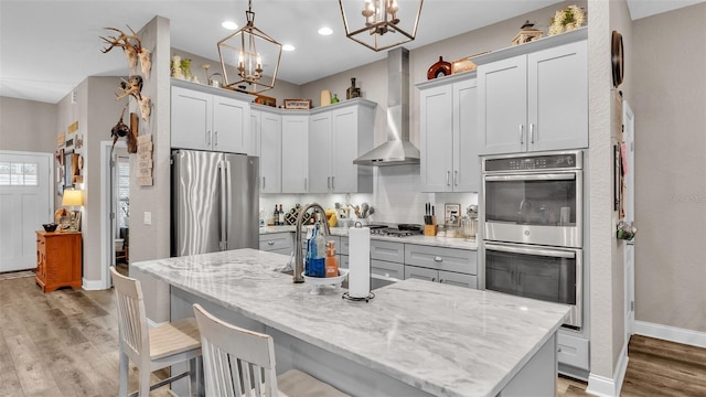 kitchen with light stone countertops, appliances with stainless steel finishes, wall chimney exhaust hood, pendant lighting, and an island with sink