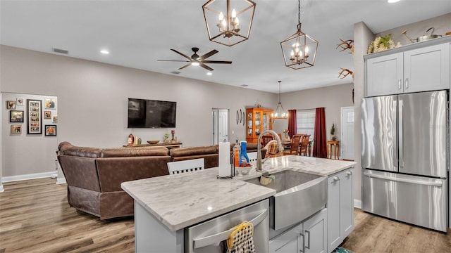 kitchen featuring a kitchen island with sink, ceiling fan with notable chandelier, stainless steel fridge, decorative light fixtures, and light stone counters