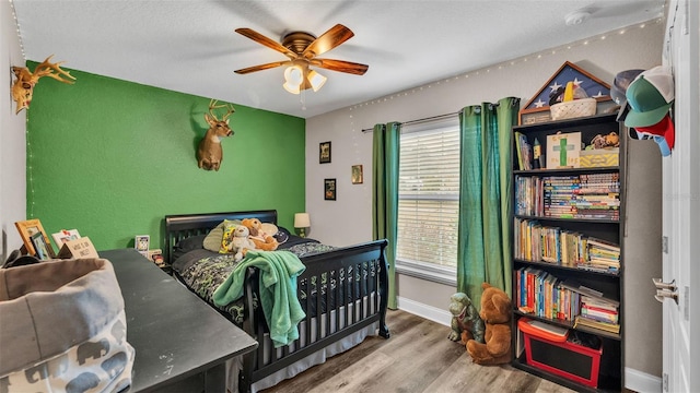 bedroom with wood-type flooring, a nursery area, and ceiling fan