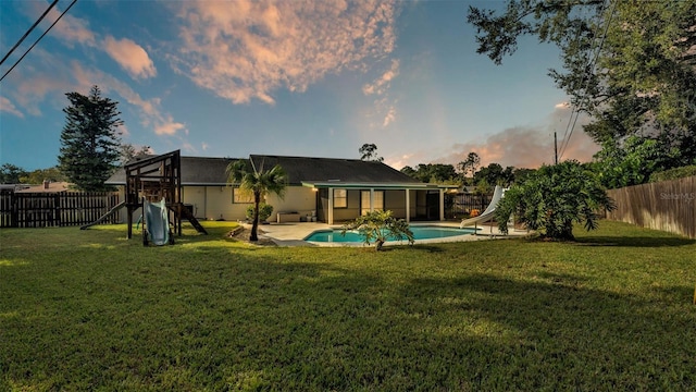 back house at dusk with a lawn, a fenced in pool, and a playground