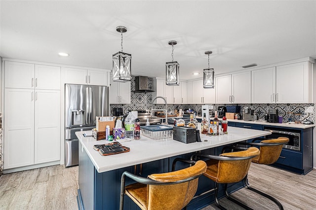 kitchen featuring appliances with stainless steel finishes, a kitchen island with sink, hanging light fixtures, wall chimney range hood, and white cabinets