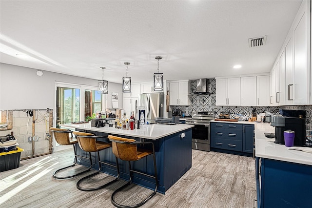 kitchen featuring an island with sink, stainless steel appliances, wall chimney range hood, pendant lighting, and white cabinets