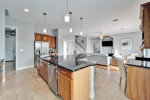 kitchen featuring sink, appliances with stainless steel finishes, an island with sink, decorative light fixtures, and dark stone counters