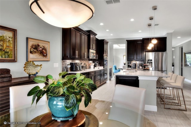 kitchen featuring an island with sink, appliances with stainless steel finishes, decorative backsplash, decorative light fixtures, and dark brown cabinets