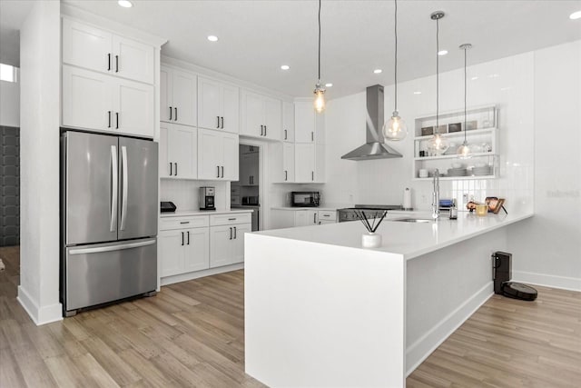 kitchen featuring wall chimney range hood, white cabinets, hanging light fixtures, and stainless steel refrigerator