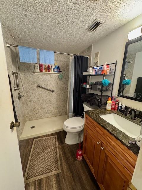 full bath featuring a textured ceiling, toilet, tiled shower, visible vents, and wood finished floors