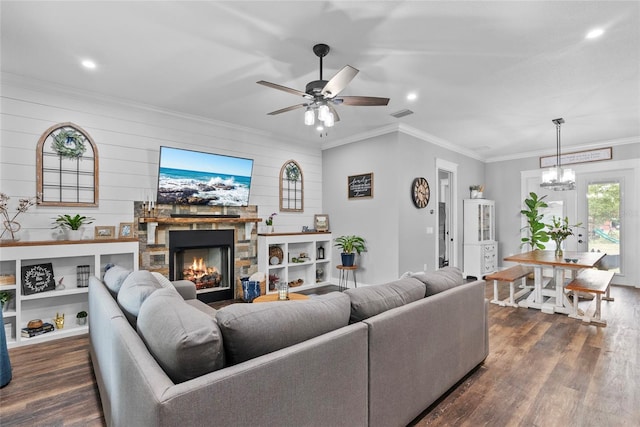 living room with ceiling fan with notable chandelier, dark wood-type flooring, crown molding, and a stone fireplace