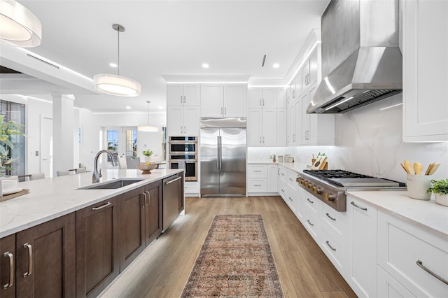 kitchen featuring hanging light fixtures, appliances with stainless steel finishes, sink, white cabinetry, and wall chimney range hood