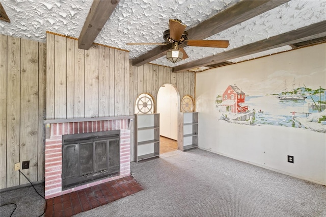 unfurnished living room with wood walls, a brick fireplace, carpet flooring, beamed ceiling, and ceiling fan