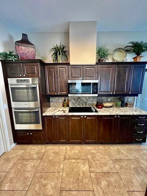 kitchen with appliances with stainless steel finishes, dark brown cabinets, light stone counters, light tile patterned flooring, and backsplash