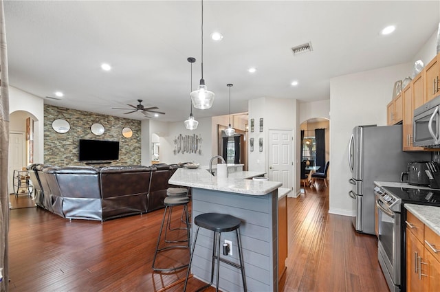 kitchen with stainless steel appliances, an island with sink, hanging light fixtures, light stone counters, and a breakfast bar