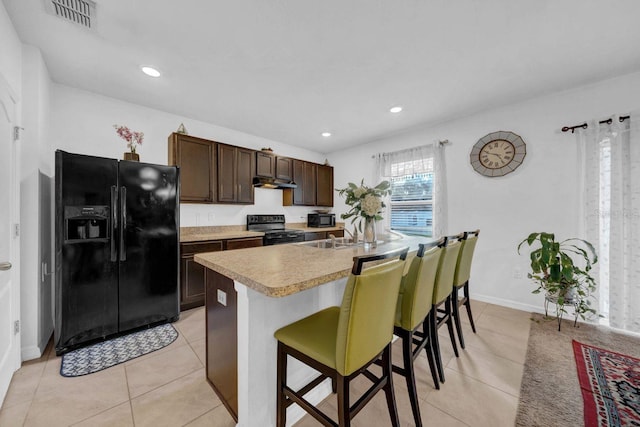 kitchen with dark brown cabinetry, black appliances, an island with sink, light tile patterned flooring, and a breakfast bar