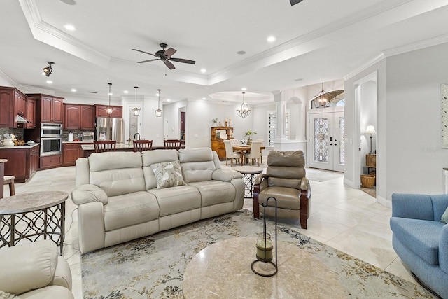 living room featuring a tray ceiling, ceiling fan with notable chandelier, ornamental molding, and decorative columns