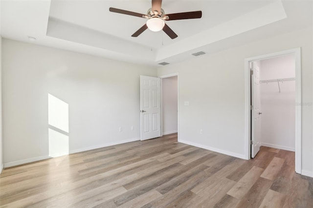 unfurnished bedroom with a spacious closet, a raised ceiling, a closet, ceiling fan, and light hardwood / wood-style floors