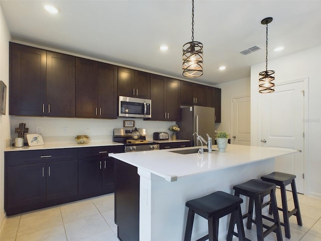 kitchen with a kitchen island with sink, stainless steel appliances, a sink, hanging light fixtures, and light countertops