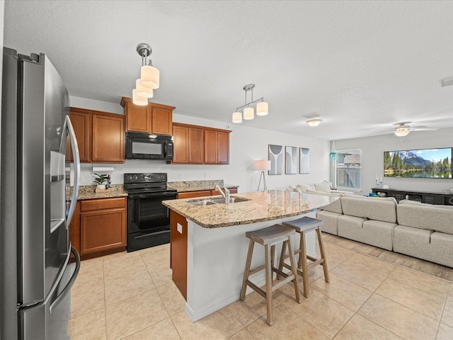 kitchen with sink, hanging light fixtures, light stone counters, black appliances, and an island with sink