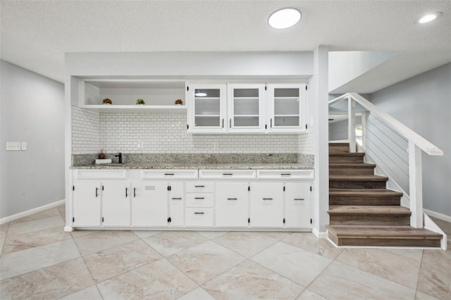 kitchen with light stone counters, open shelves, backsplash, glass insert cabinets, and white cabinets