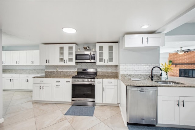 kitchen with appliances with stainless steel finishes, a ceiling fan, glass insert cabinets, a sink, and light stone countertops