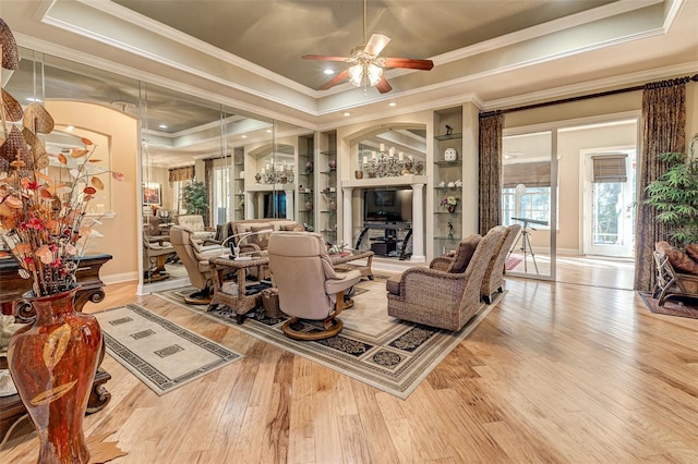 living room with a raised ceiling, crown molding, built in shelves, and light hardwood / wood-style flooring