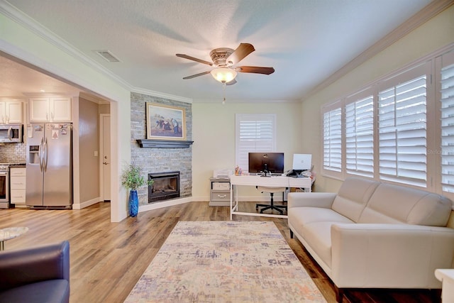 living room featuring crown molding, light hardwood / wood-style flooring, ceiling fan, a textured ceiling, and a stone fireplace