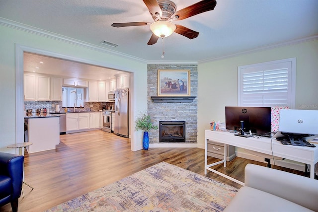 living room with crown molding, ceiling fan, a textured ceiling, a stone fireplace, and light wood-type flooring