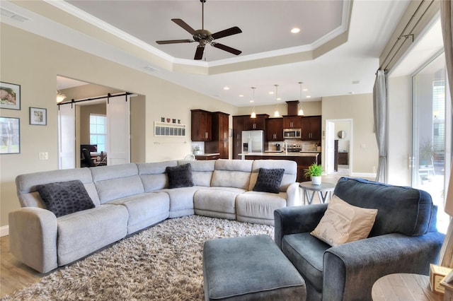 living room featuring crown molding, light hardwood / wood-style flooring, a tray ceiling, ceiling fan, and a barn door