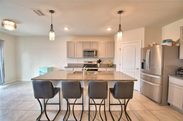 kitchen with light stone counters, decorative light fixtures, light tile patterned floors, appliances with stainless steel finishes, and an island with sink