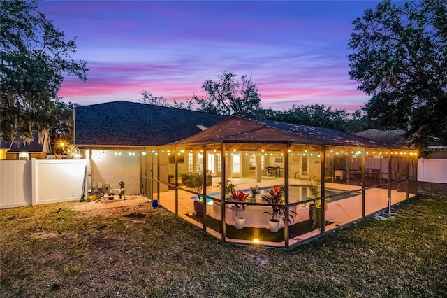 back house at dusk with a lawn, a patio, and glass enclosure