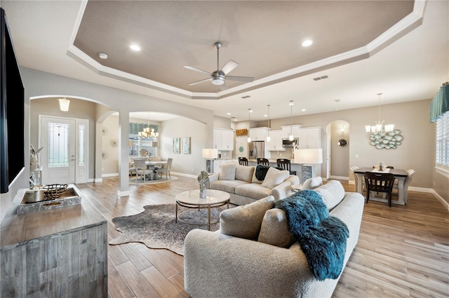 living area with ceiling fan with notable chandelier, baseboards, arched walkways, light wood-style floors, and a raised ceiling