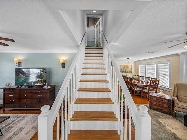 stairway with ceiling fan, ornamental molding, wood-type flooring, and a textured ceiling