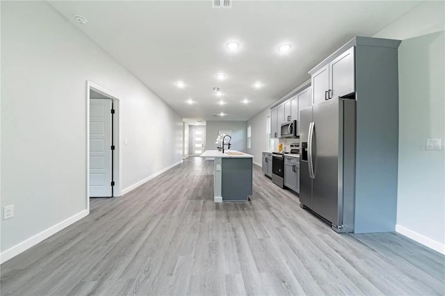 kitchen with light wood-type flooring, a kitchen island with sink, a sink, appliances with stainless steel finishes, and light countertops