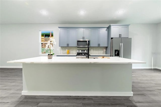 kitchen with wood finished floors, a center island with sink, light countertops, appliances with stainless steel finishes, and backsplash