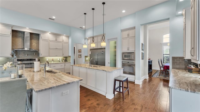 kitchen with pendant lighting, sink, built in appliances, an island with sink, and wall chimney exhaust hood