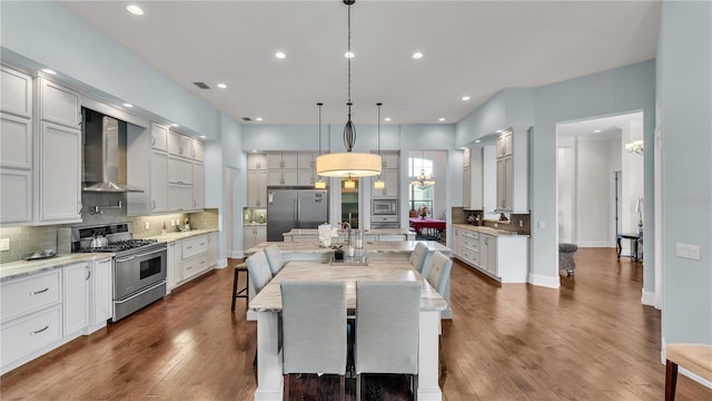 kitchen with wall chimney exhaust hood, decorative light fixtures, stainless steel appliances, a large island, and light stone countertops