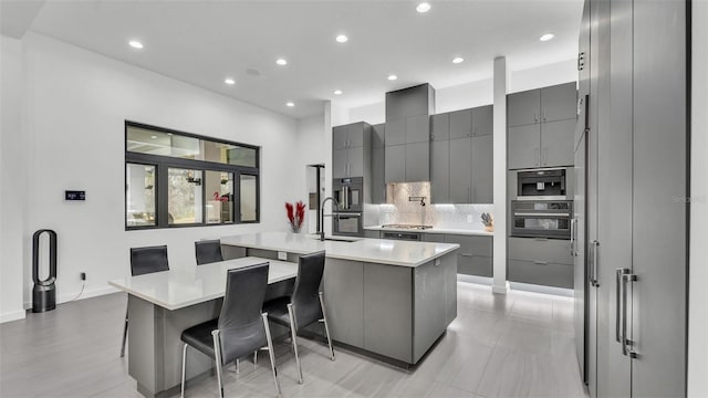 kitchen with gray cabinetry, a kitchen breakfast bar, black double oven, tasteful backsplash, and an island with sink