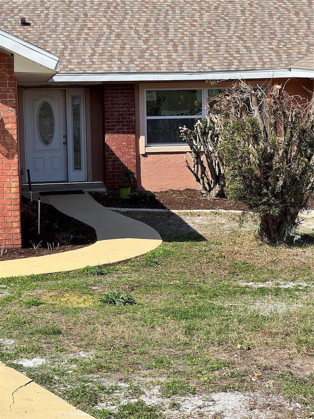 view of exterior entry featuring a shingled roof, brick siding, and a yard