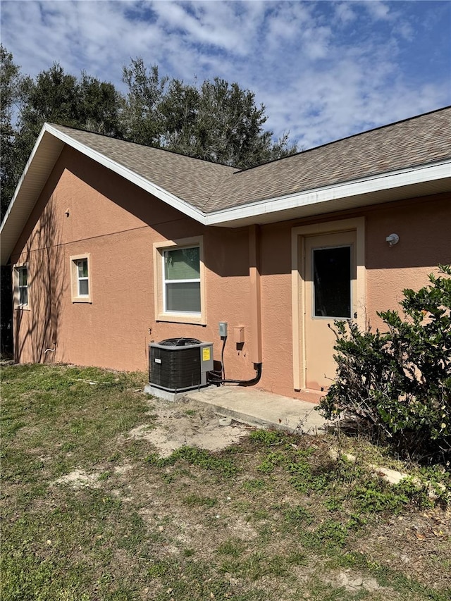 rear view of house with a shingled roof, stucco siding, and central AC unit