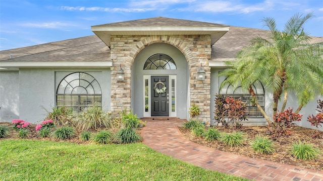 entrance to property featuring a yard, a shingled roof, stone siding, and stucco siding