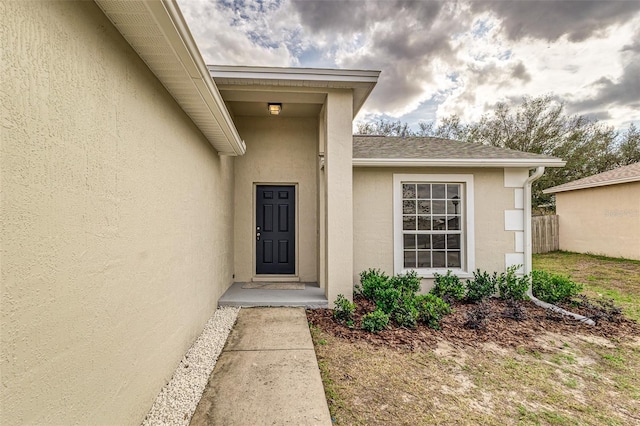 entrance to property featuring a shingled roof, fence, and stucco siding