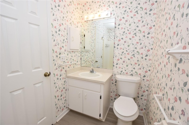 bathroom with toilet, wallpapered walls, wood finished floors, and vanity