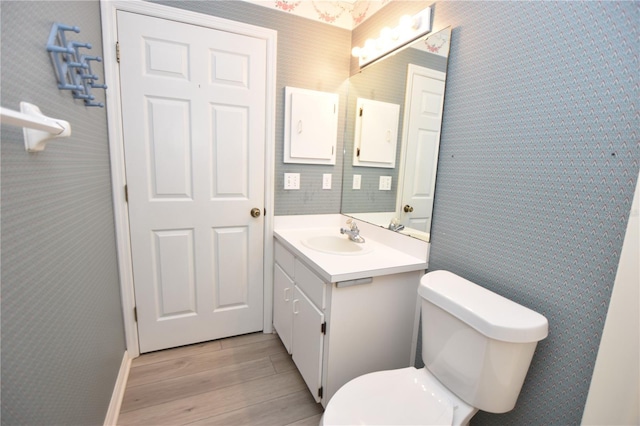 half bathroom with toilet, wallpapered walls, wood finished floors, and vanity