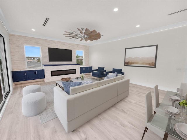 living area with visible vents, a ceiling fan, a glass covered fireplace, light wood-style flooring, and ornamental molding