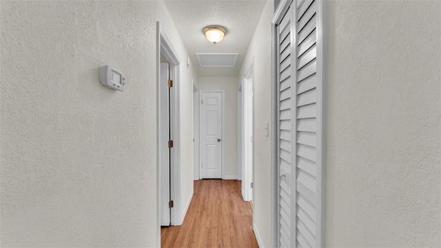 hall featuring light wood finished floors, attic access, a textured ceiling, and a textured wall