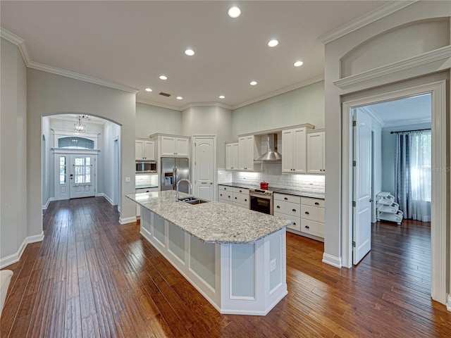 kitchen featuring a sink, appliances with stainless steel finishes, arched walkways, wall chimney exhaust hood, and a kitchen island with sink